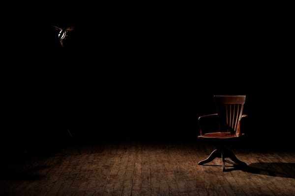 An empty wooden chair sits in the center of a spot light against a black background.